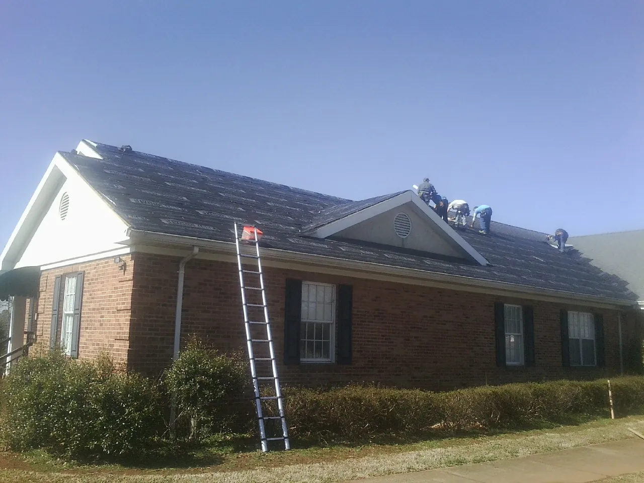 Crew tearing shingles off a brick ranch roof with a ladder set — Emergency Roof Repair work by our crew in Petal