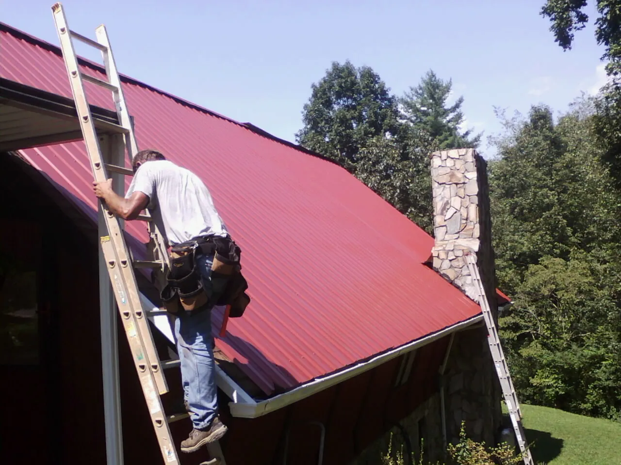 Roofer on a ladder beside a red metal roof and stone chimney — Soffit & Fascia Repair by Roofing Brothers in Petal